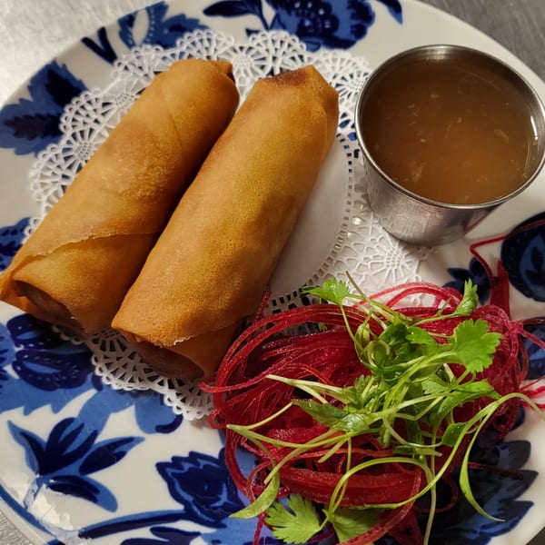 Two golden-brown Thai spring rolls with dipping sauce and a side of red and green garnish.