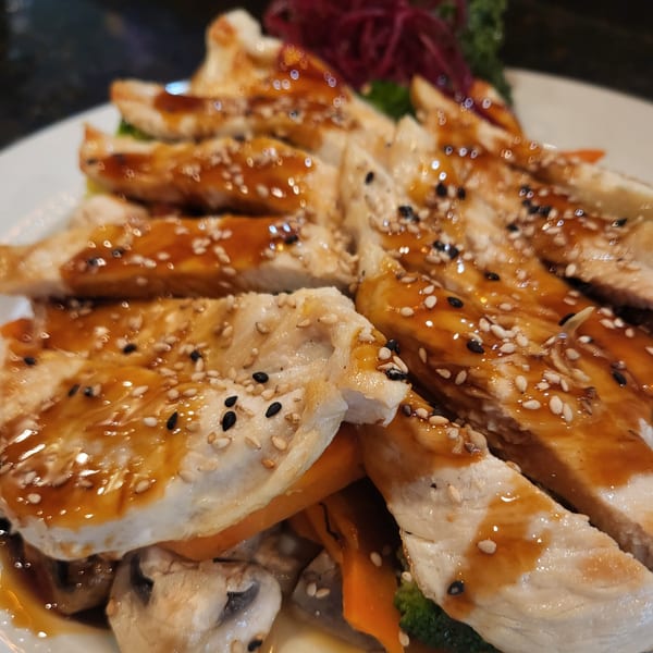 Sliced chicken breast with a glossy brown sauce, white and black sesame seeds, and vegetables.