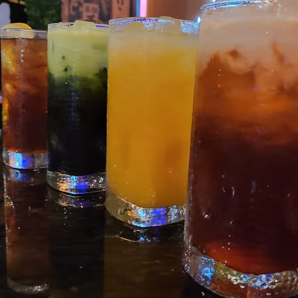 Four colorful Thai drinks, including iced tea and green tea, in tall glasses on a dark surface.